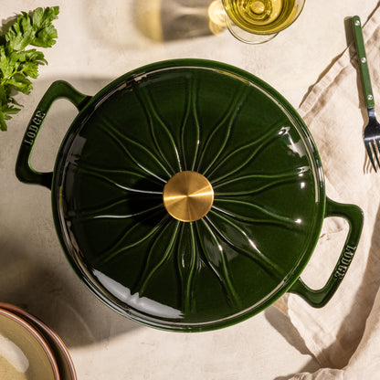 A dark green Lodge enameled cast iron pot with a lid, featuring a starburst pattern and a gold-tone knob, shown from a top-down angle on a beige linen surface with dining props.