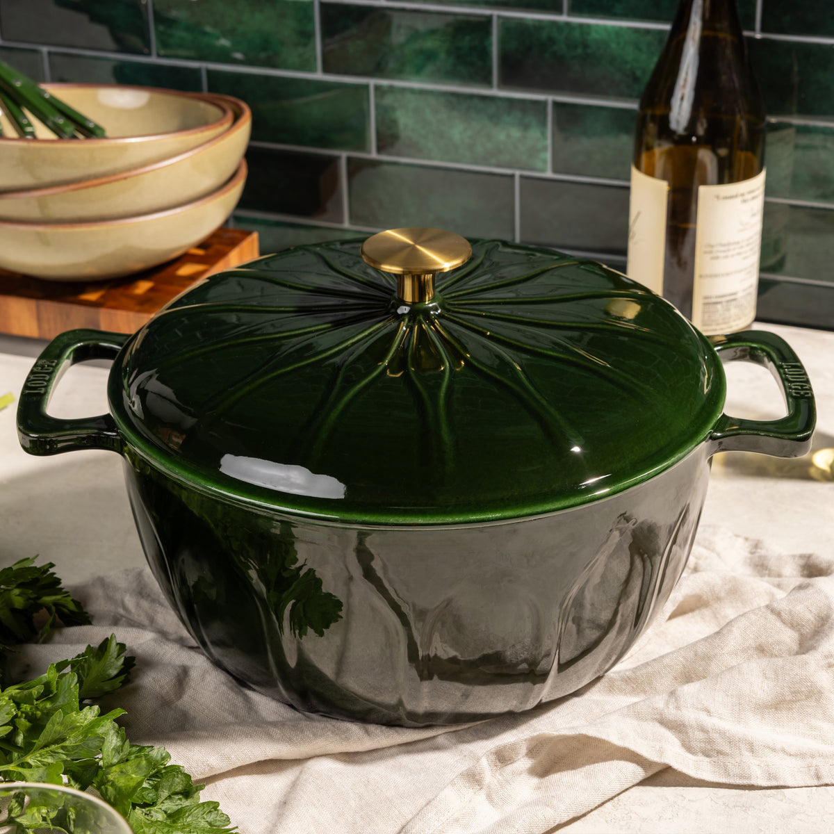 A glossy dark green enameled cast iron Dutch oven with a matching lid and gold knob, styled on a beige cloth with kitchen props like a bottle, bowls, and parsley in the background.