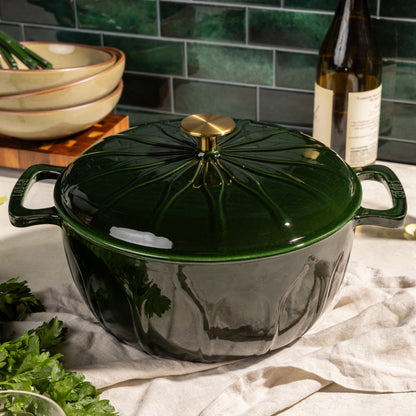 A glossy dark green enameled cast iron Dutch oven with a matching lid and gold knob, styled on a beige cloth with kitchen props like a bottle, bowls, and parsley in the background.