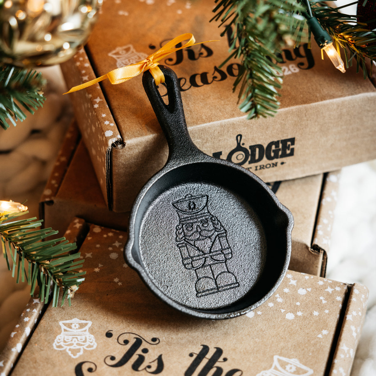 A small black cast-iron nutcracker ornament shaped like a miniature skillet, displayed on top of brown gift boxes with holiday greenery and warm string lights around it.