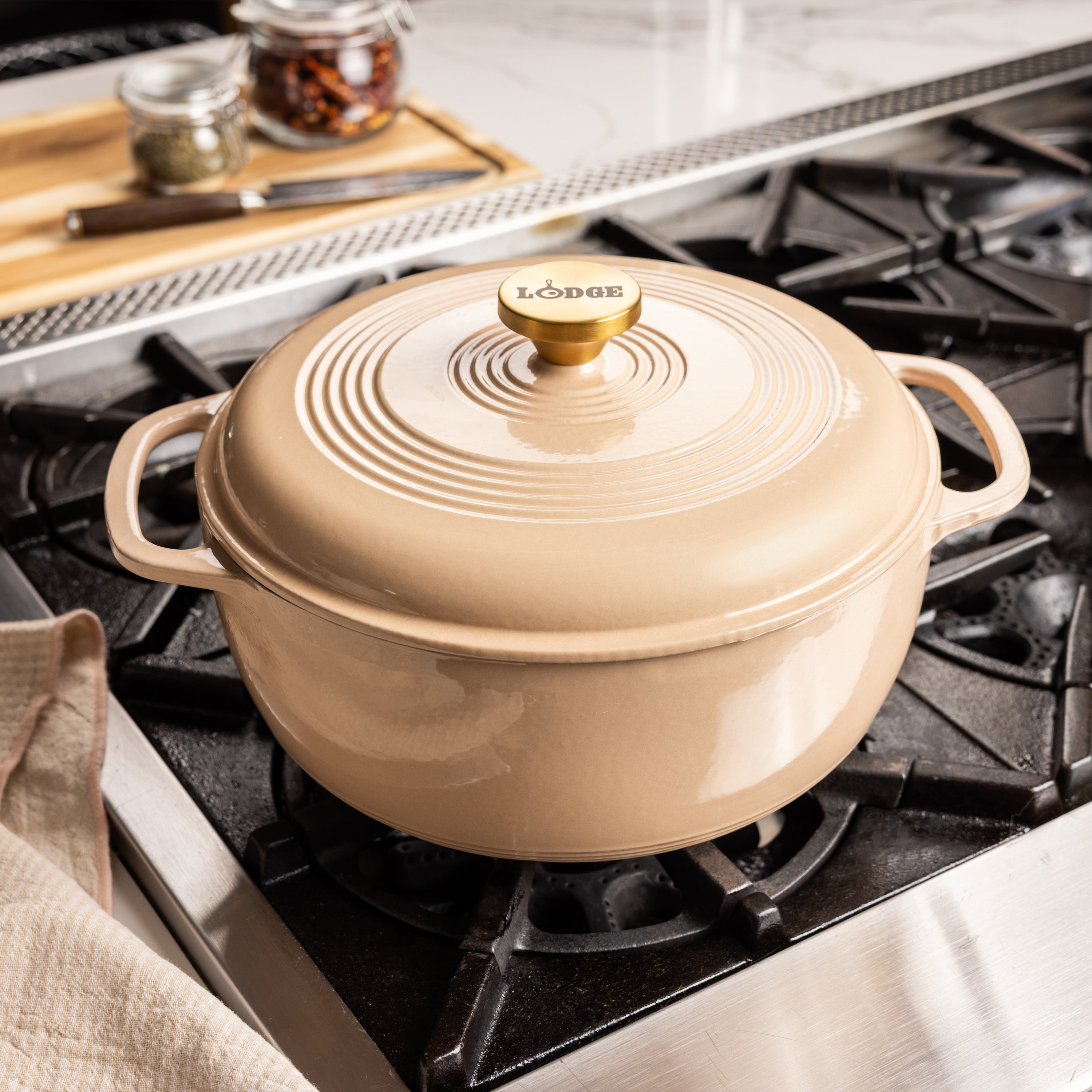 A Lodge enameled cast iron Dutch oven with lid in a warm beige color, sitting on a black gas stovetop in a kitchen setting.