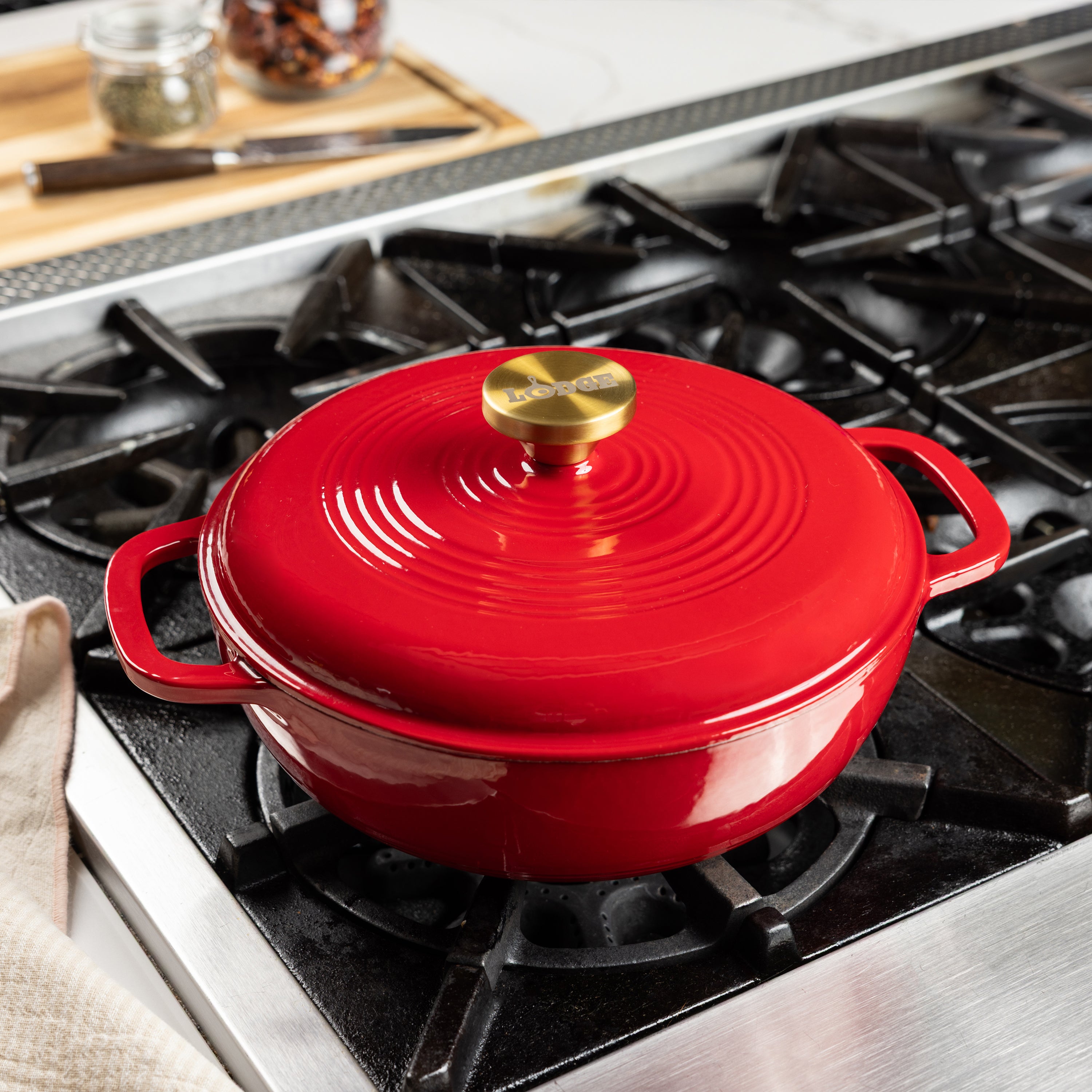 A red enameled cast iron Dutch oven with a matching lid and gold-tone knob, sitting on a black gas stovetop in a kitchen setting.