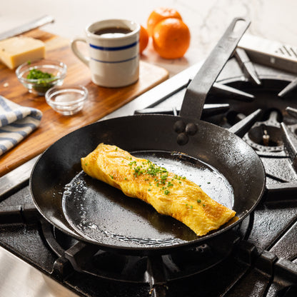 A black carbon steel skillet on a gas stovetop, cooking an omelet with green herb garnish.