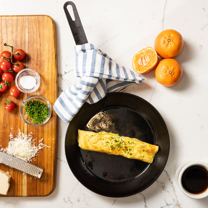 A carbon steel skillet with a freshly prepared omelet, styled in a bright kitchen flat-lay with tomatoes, oranges, cheese, and coffee.