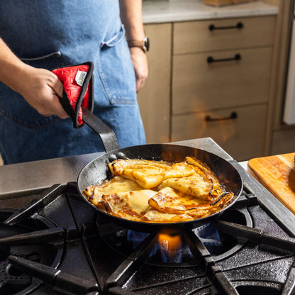 A person in a blue apron holds a carbon steel skillet while cooking crepes on a gas stove burner in a kitchen.