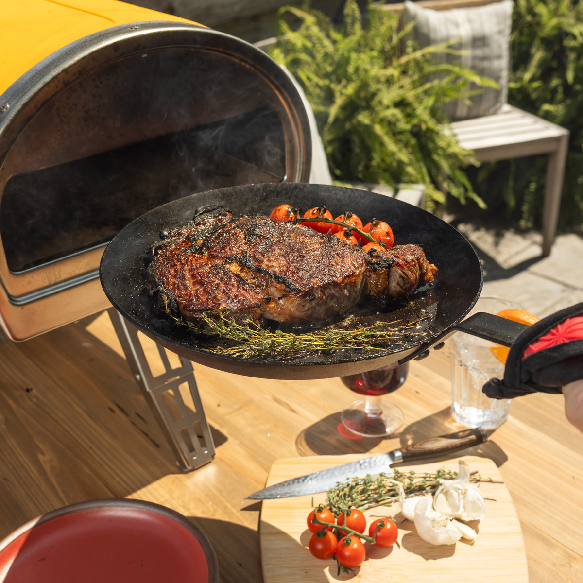 Someone removing skillet from an outdoor  pizza oven, revealing a perfectly seared steak.