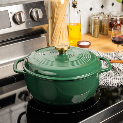 A green enameled cast iron Dutch oven with a matching lid sits on a black induction stovetop in a kitchen setting. The pot has side handles and a gold-tone knob on the lid, with a checkered cloth under the pot and kitchen items like spaghetti, oil, and spices in the background.