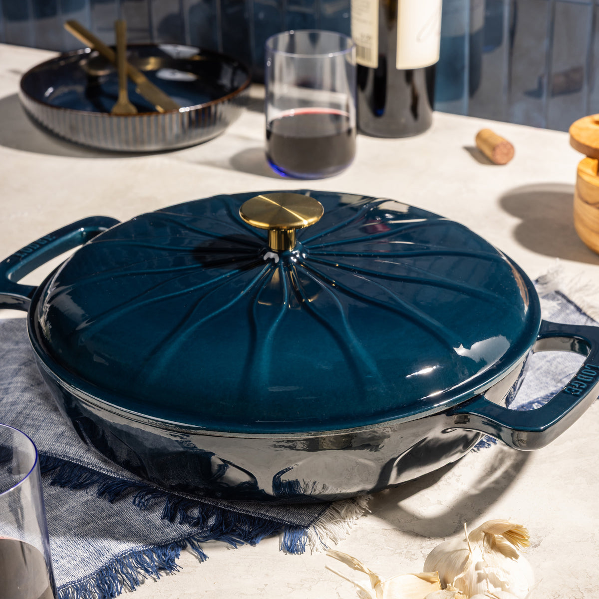 A dark teal enameled cast iron pot with a matching lid and a gold-tone knob, styled on a table with a glass of red wine, garlic, and dining props in the background.