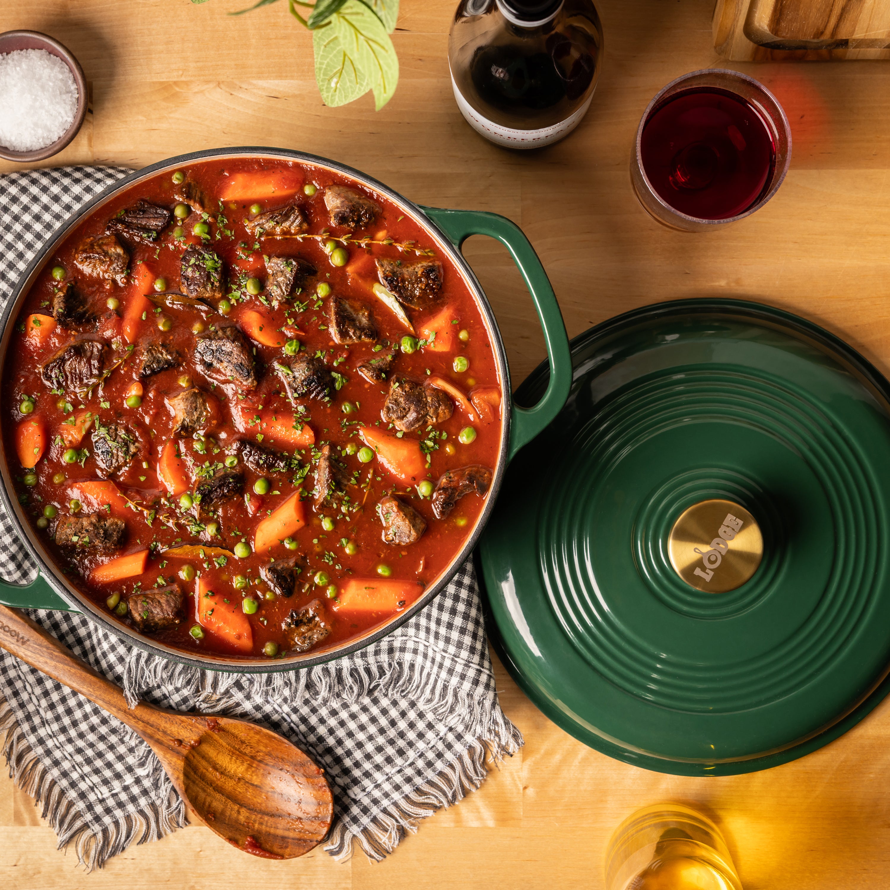 A green enameled cast iron Dutch oven filled with a hearty beef and carrot stew, shown on a wooden table alongside its matching lid, a wooden spoon, a checkered cloth, and dining props.