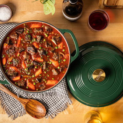 A green enameled cast iron Dutch oven filled with a hearty beef and carrot stew, shown on a wooden table alongside its matching lid, a wooden spoon, a checkered cloth, and dining props.
