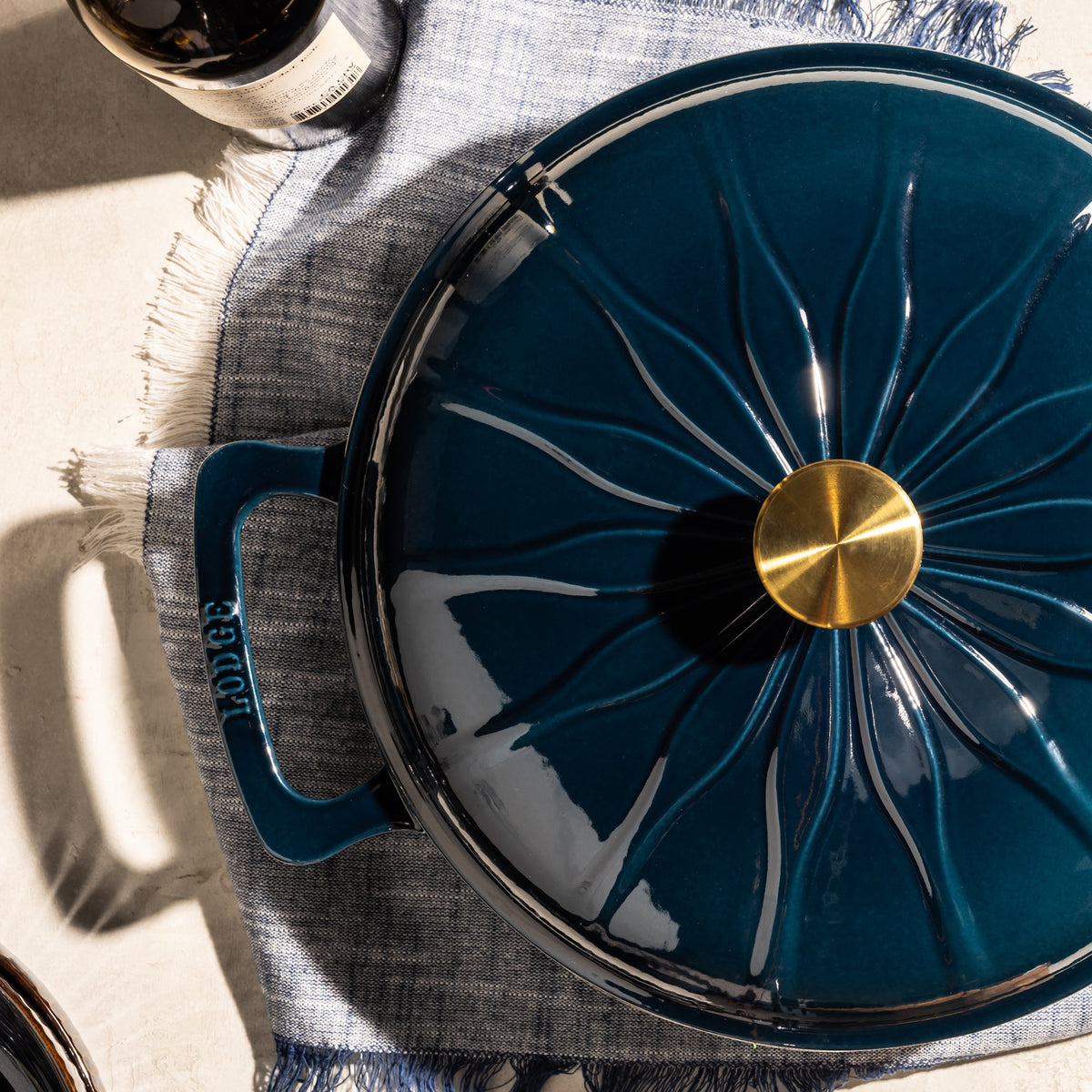A dark teal blue enameled cast iron Dutch oven with a matching lid featuring a starburst pattern and a gold knob, styled on a blue fringed placemat with a bottle partially visible.