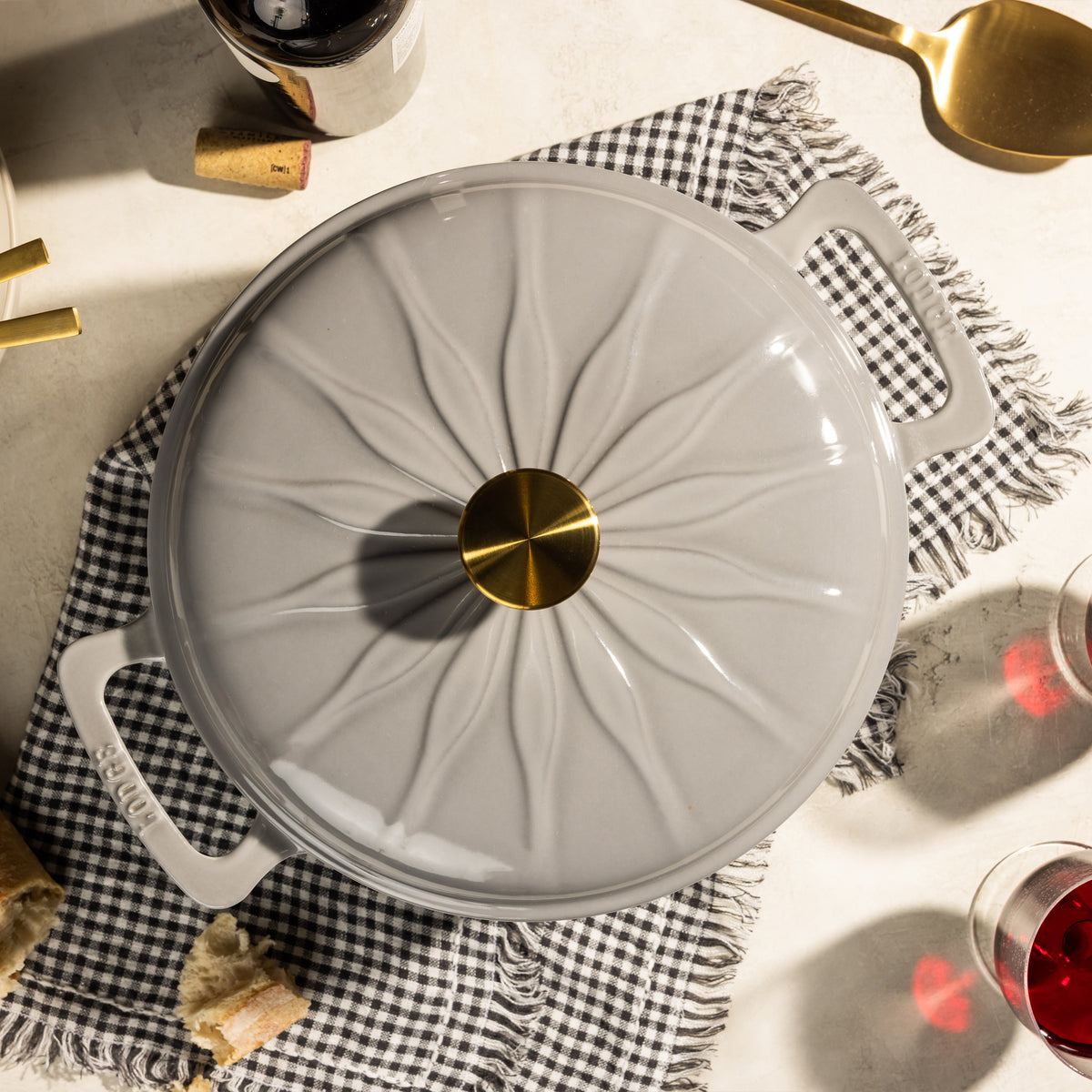 A light gray enameled cast iron pot with a lid featuring a sunburst pattern and a shiny gold knob, styled on a black and white checkered napkin with wine glasses, corks, and a gold spoon.