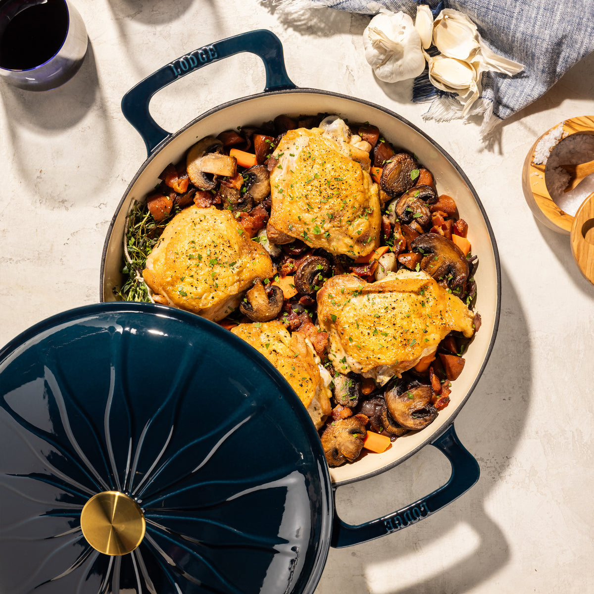 A dark blue enameled cast iron Dutch oven with a matching lid, shown filled with roasted chicken, mushrooms, and vegetables on a light stone surface. The lid is partially lifted in the foreground, and garlic, a glass of red wine, and wooden salt and pepper mills are arranged around it.