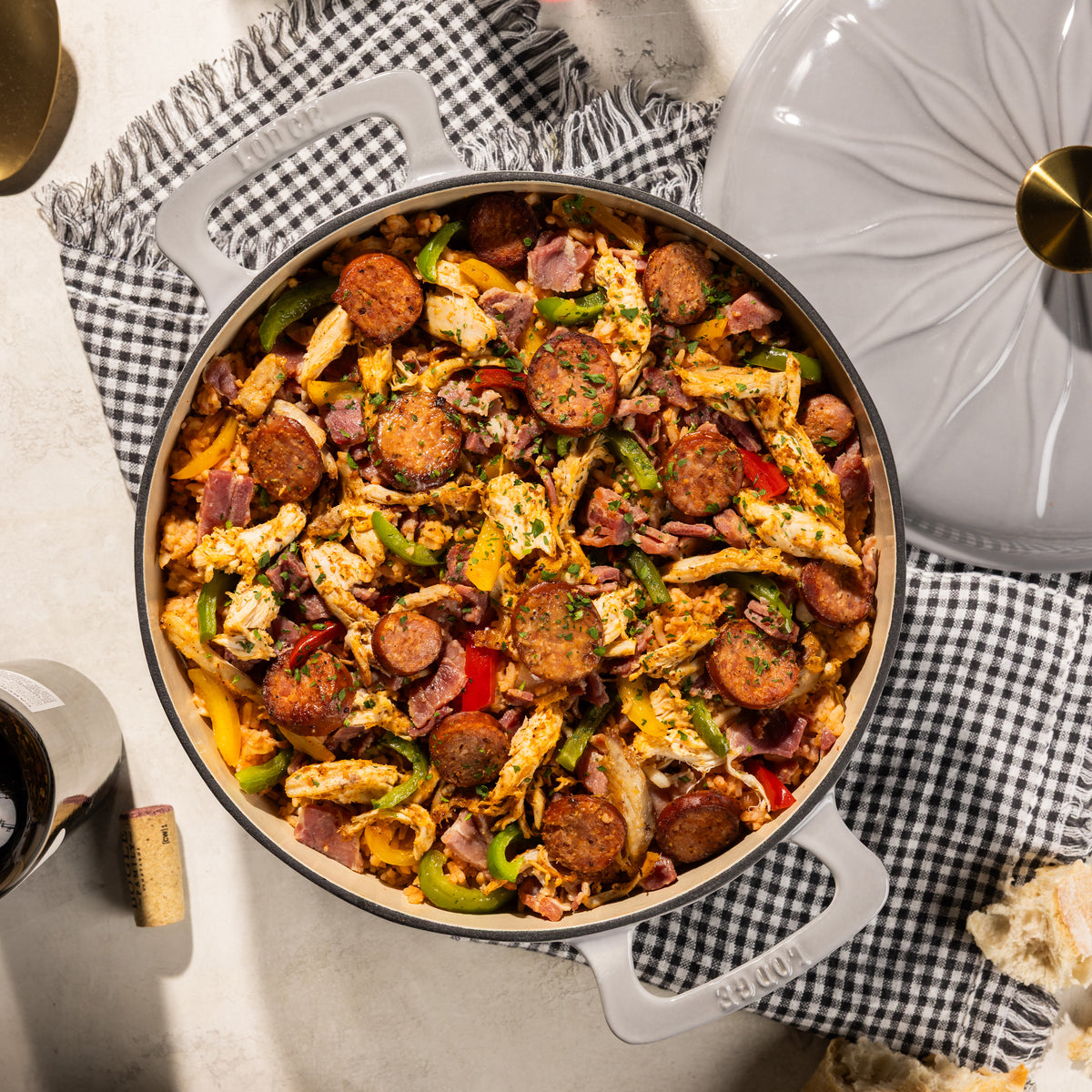 An overhead shot of a cast iron Dutch oven filled with a cooked sausage and chicken dish, placed on a black-and-white checkered cloth next to the lid on a light stone surface.