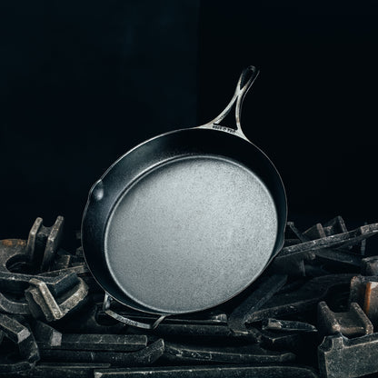 A dark cast iron skillet with a textured non-stick surface, resting on a pile of dark metal tools against a solid black background.