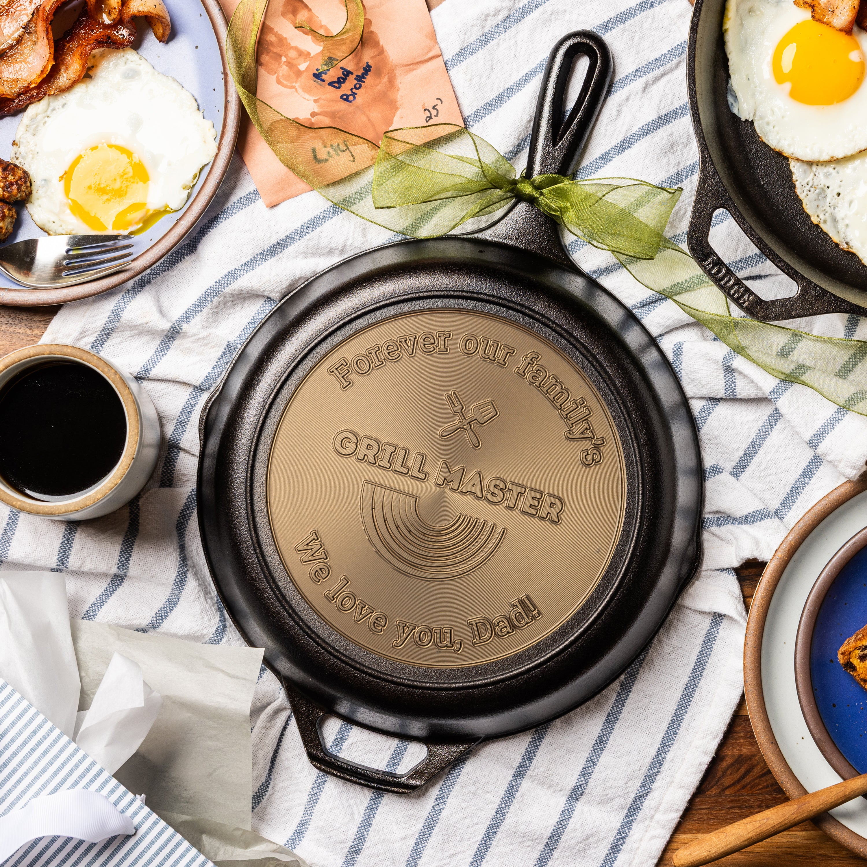 Engraved personalized skillet displayed on a breakfast table as a gift for Dad, featuring “forever our family's” along the top arc, “grill master” in the center, and “we love you, dad!” along the bottom arc.