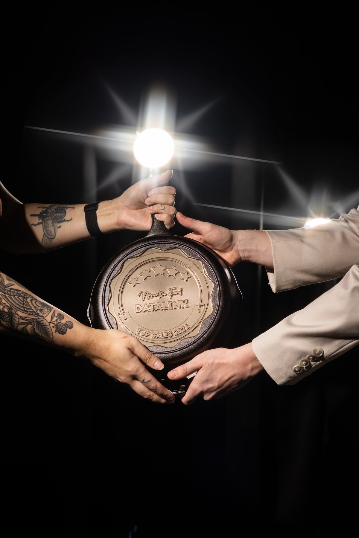 Three hands holding a round object with a light source above, on a dark background