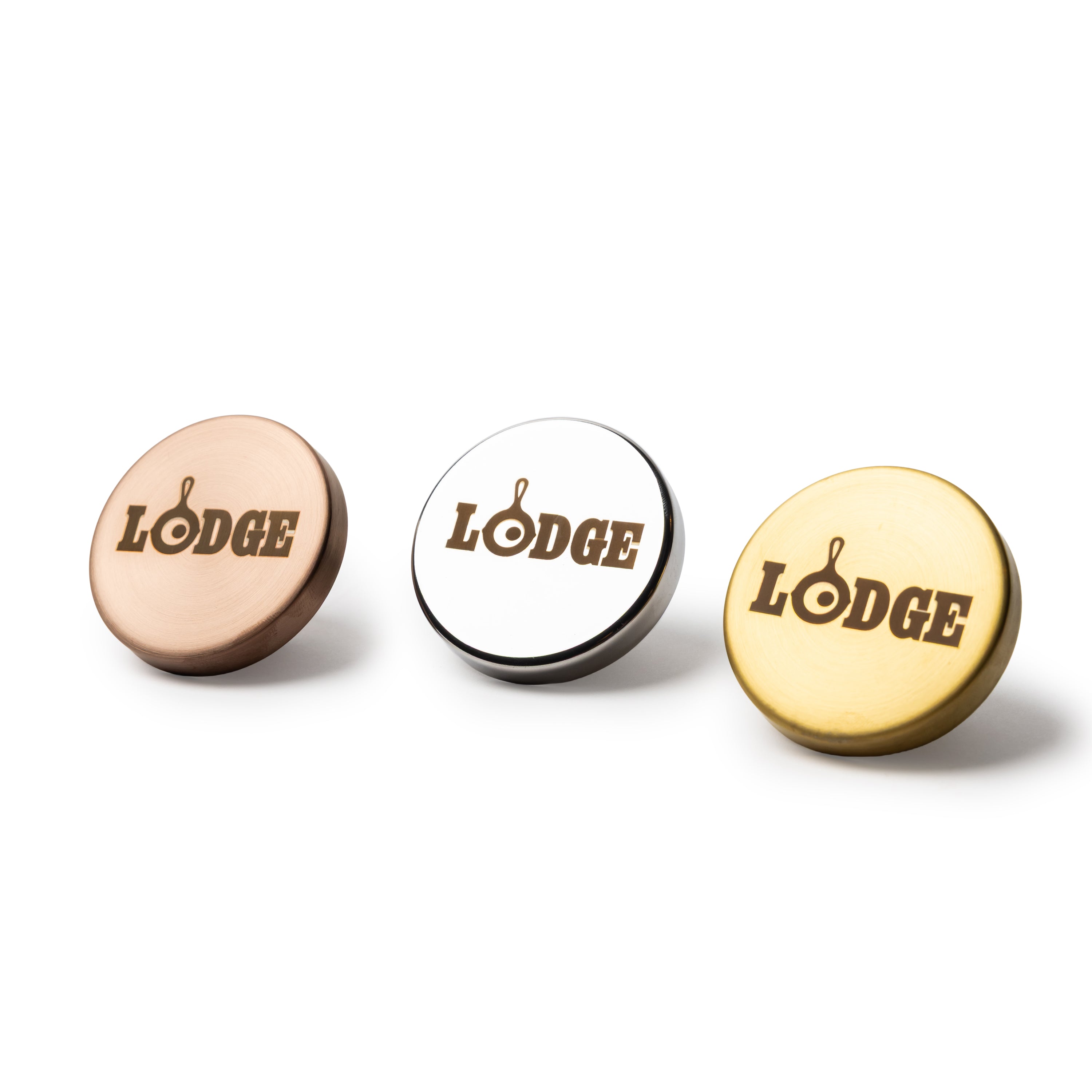 Three round metal dutch oven knobs with the word 'LODGE' printed in brown on the top, shown in rose gold, silver, and gold finishes on a white background.