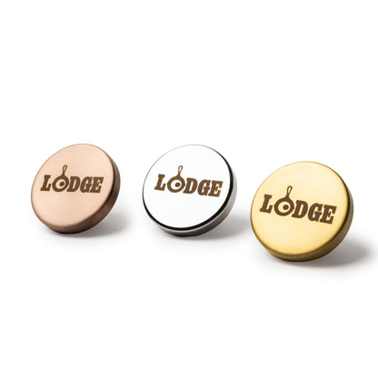 Three round metal dutch oven knobs with the word 'LODGE' printed in brown on the top, shown in rose gold, silver, and gold finishes on a white background.