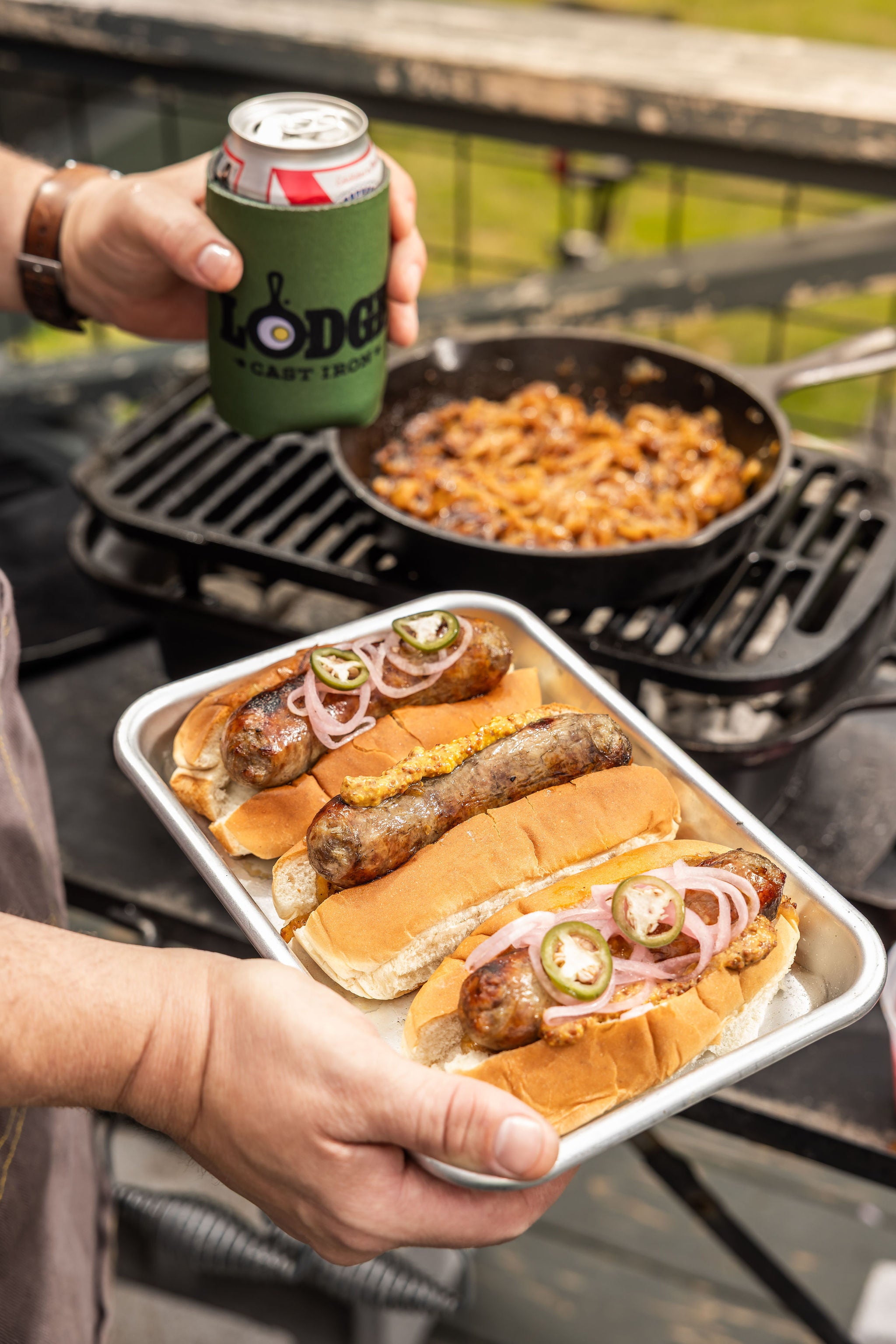 Person holding a tray of hot dogs with buns and sauerkraut, standing next to a grill with food cooking.