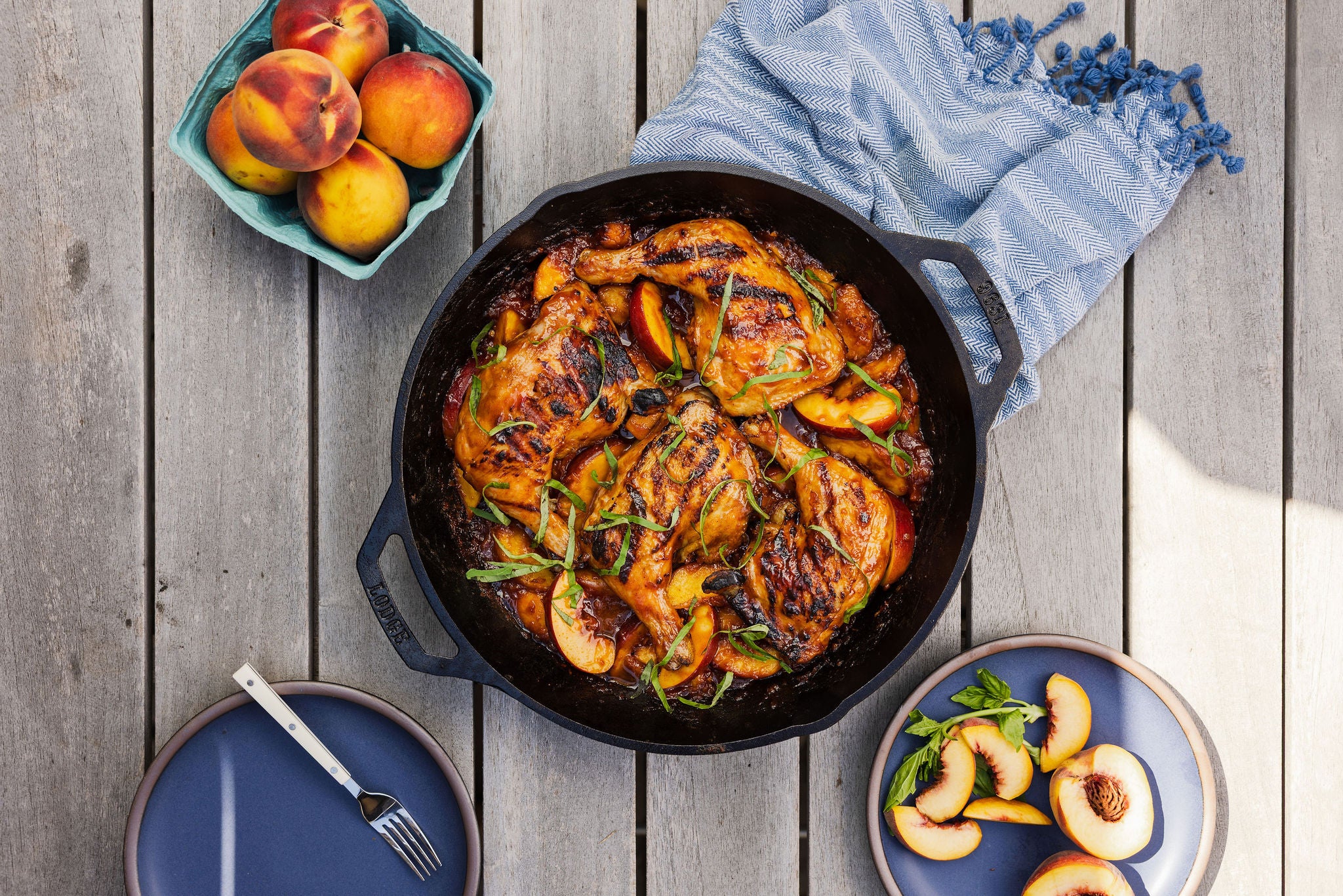 Peach barbecue chicken in a cast iron dual handle pan surrounded by sliced and whole peaches on a table