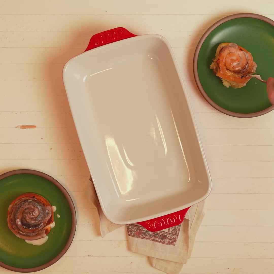 Fresh cinnamon rolls beside cherry-red stoneware casserole dish.
